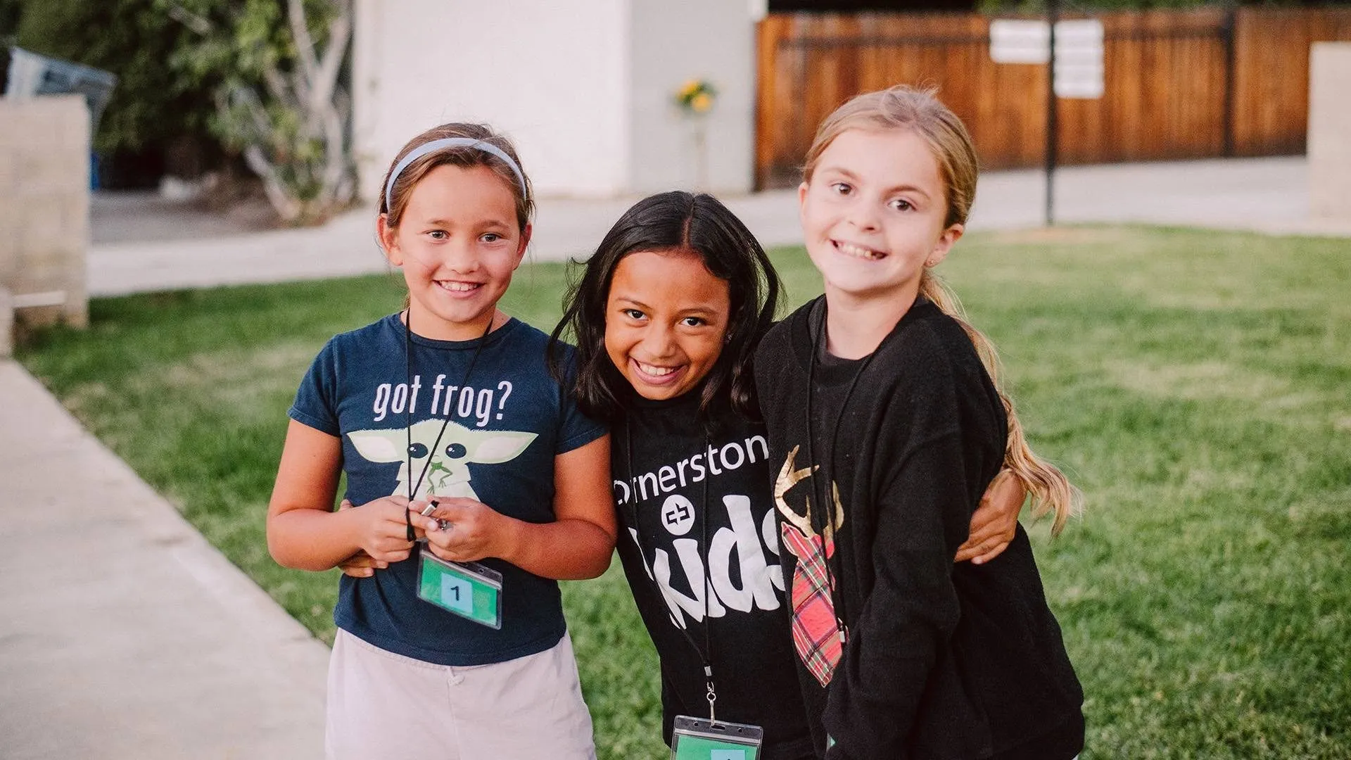 Three smiling elementary-age girls with their arms around each other on the lawn